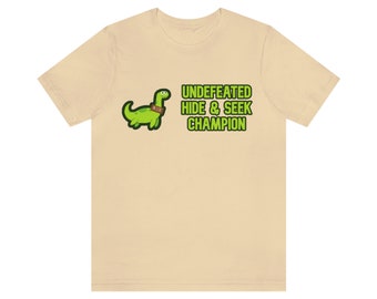 May include: A beige t-shirt with a green cartoon dinosaur wearing a brown tag that says "HIDER" and the text "UNDEFEATED HIDE & SEEK CHAMPION" in green.