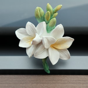 May include: A white ceramic flower air freshener with green leaves and buds. The flower is a stylised version of a freesia.