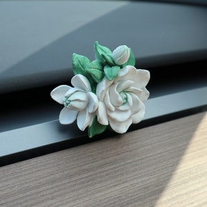 May include: A white ceramic car air freshener shaped like a bouquet of gardenias with green leaves.