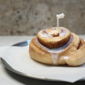 May include: A cinnamon roll candle with white icing and a small white candle wick. The candle sits on a white paper on a silver plate. The background is a neutral color.