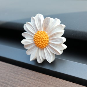 May include: A white daisy flower with a bright orange centre. The flower has multiple white petals and is set against a dark background. The daisy is a decorative item, possibly a brooch or a decorative accent.