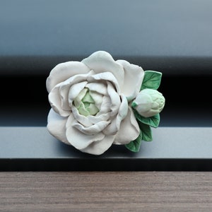 May include: A white porcelain flower brooch with green leaves. The flower has a bud and is in bloom.