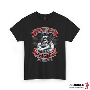 Peut inclure: T-shirt noir avec un graphisme d'un homme moustachu tenant une clé à molette. Le texte en rouge et blanc indique "Alles Wird Gut" et "Der Meister Ist Jetzt Da". Le logo de Required Clothing Company est en bas à droite.