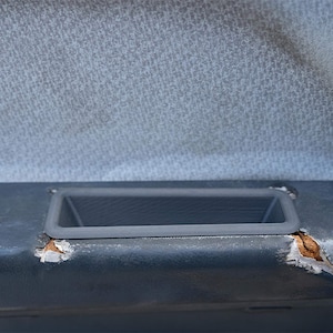 May include: A close-up of a gray fabric car door panel with a rectangular storage compartment. The fabric is worn and has a small tear.