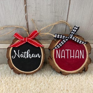 May include: Two wooden ornaments with the name "Nathan" written in white. One ornament has a black background and a red and white ribbon. The other has a red background and a black and white checkered ribbon.