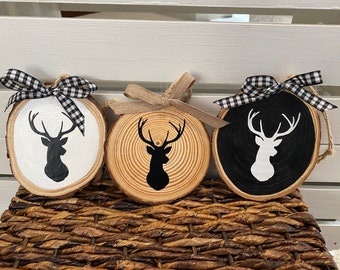 May include: Three wooden ornaments with black and white deer silhouettes. Each ornament is tied with a ribbon, one black and white checkered, one burlap, and one black and white checkered.