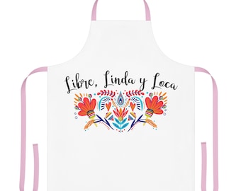 May include: A white apron with light pink neck and waist straps. The centre features the black text "Libre, Linda y Loca" above a colourful graphic of stylised flowers, leaves, and hearts in shades of red, orange, blue, and yellow.