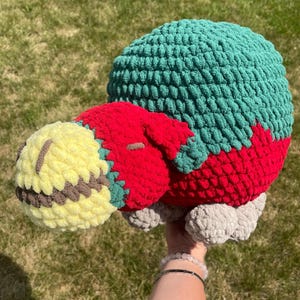 May include: A crocheted plush toy shaped like a turtle with a yellow head, red body, green shell, and gray feet.