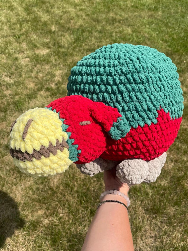 May include: A crocheted plush toy shaped like a turtle with a yellow head, red body, green shell, and gray feet.