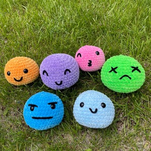 May include: A collection of seven crocheted emoji balls in various colors, including orange, purple, pink, green, blue, and light blue. Each ball features a different facial expression, such as a smile, winking, or a frown. The balls are displayed on a bed of green grass.