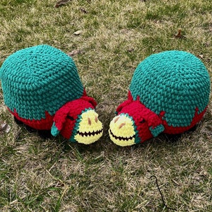 May include: Two crocheted Minecraft Sniffer plushies. The plushies are green, red, and yellow. They are sitting on a bed of green grass.