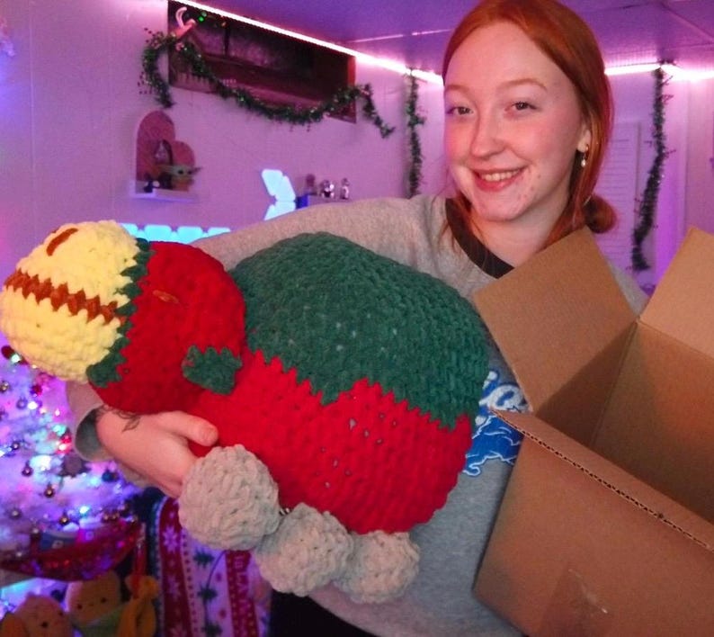 May include: A person holding a large crocheted stuffed toy shaped like a strawberry. The toy is red and green with a yellow and brown top. The person is smiling and holding a cardboard box.