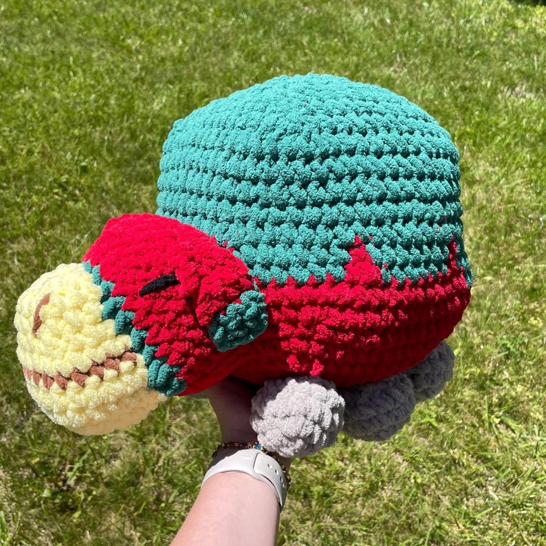 May include: A crocheted stuffed animal shaped like a dinosaur. The dinosaur is green, red, and yellow. It has a long neck and a small head with a closed eye. The dinosaur has a fluffy texture.