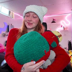 May include: A woman wearing a white crocheted hat is holding a green and red crocheted plush toy. The toy has a gray and yellow face.