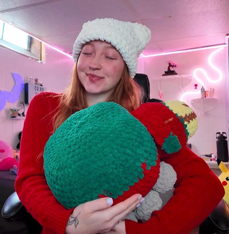 May include: A woman wearing a white crocheted hat is holding a green and red crocheted plush toy. The toy has a gray and yellow face.