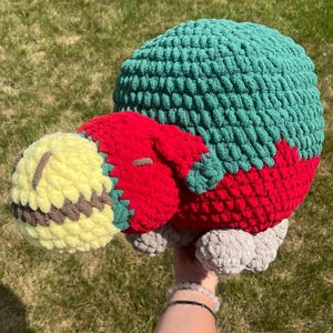 May include: A crocheted stuffed animal with a green, red, and yellow body. The animal has a round head and a long, thin body. The animal is lying on its side.