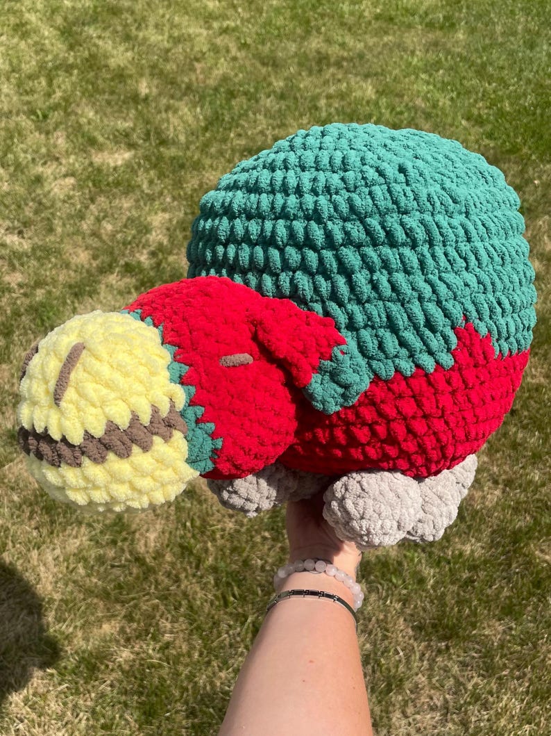 May include: A crocheted stuffed animal with a green, red, and yellow body. The animal has a round head and a long, thin body. The animal is lying on its side.