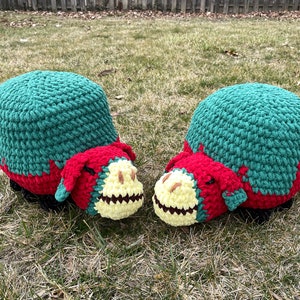 May include: Two crocheted dinosaur-shaped footstools in green, red, and yellow. The footstools are facing each other and are sitting on a patch of green grass.