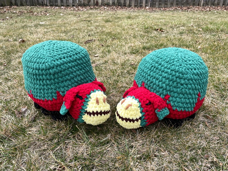 May include: Two crocheted dinosaur-shaped footstools in green, red, and yellow. The footstools are facing each other and are sitting on a patch of green grass.
