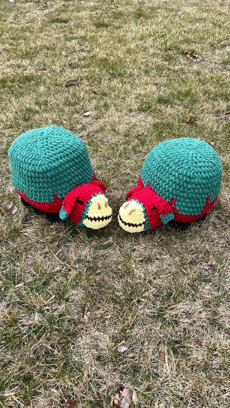 May include: Two crocheted plush toys shaped like a cartoon animal with a green, red, and yellow color scheme. The toys are sitting on a bed of green grass.
