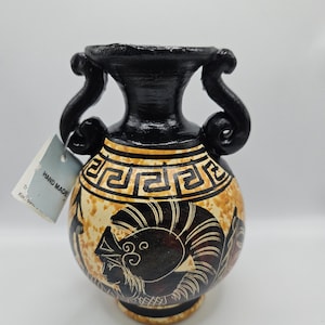 Peut inclure: Un vase en céramique noir et blanc avec un motif de clé grecque et une représentation d'un guerrier portant un casque. Le vase est fait à la main et possède deux anses.