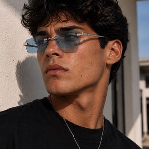 May include: Rimless sunglasses with blue tinted lenses and silver frames. The sunglasses have a rectangular shape and are worn by a person. A silver cross pendant hangs from a chain around the person's neck.