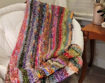 May include: A colorful striped crocheted throw blanket draped over a white armchair. The blanket is made of various shades of red, green, blue, purple, and brown yarn.