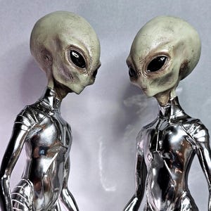 May include: Two alien figures with large black eyes and gray skin stand facing each other. They wear shiny silver suits. The figures have elongated heads and slender necks, creating a futuristic and otherworldly appearance.