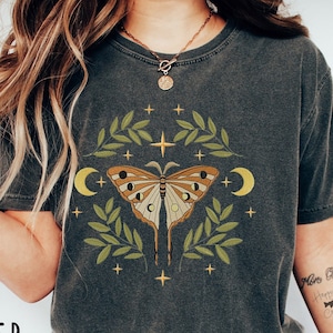 May include: A dark gray t-shirt with a graphic of a butterfly with moon and star accents. The butterfly is surrounded by green leaves and the design is centered on the shirt.