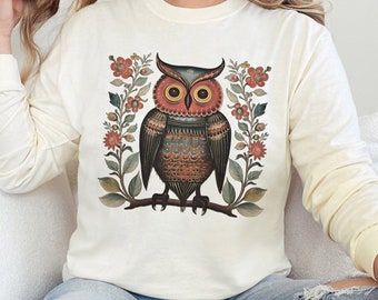 May include: A cream colored long-sleeved sweatshirt with a graphic of a brown and blue owl with red eyes surrounded by red and green flowers. The text "IVORY" is in the upper right corner of the image.