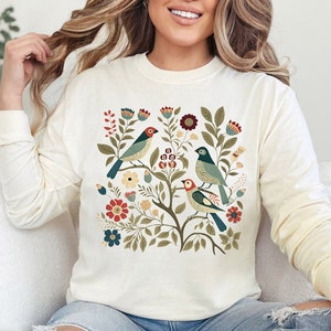 May include: A cream-colored long-sleeved sweatshirt with a floral design featuring blue and green birds. The design includes red, green, and yellow flowers.