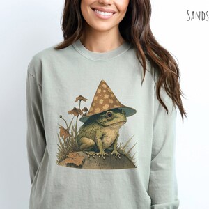 Pode incluir: Um sapo verde claro usando um chap&eacute;u marrom e branco em forma de cogumelo com bolinhas brancas est&aacute; sentado em uma rocha cercada por folhagem verde. O sapo est&aacute; usando um chap&eacute;u com uma ponta pontiaguda e uma aba larga. O sapo est&aacute; sentado em uma rocha com uma superf&iacute;cie marrom. O sapo est&aacute; cercado por folhagem verde. O fundo &eacute; de cor verde claro.