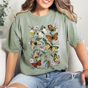May include: A green t-shirt featuring a vintage-style print of butterflies and flowers. The butterflies are in various colours, including orange, yellow, blue, and red.