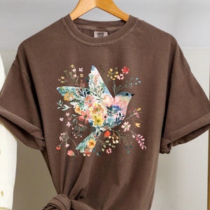 Op de afbeelding: Een bruin T-shirt met een bloemenprint met een vogel. De vogel is gemaakt van bloemen en heeft een blauwe kop en vleugels. Het shirt hangt aan een houten hanger.
