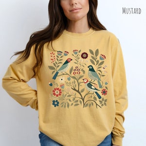 May include: A mustard yellow long-sleeved shirt with a floral and bird design. The design features three birds with blue, green, and red accents, surrounded by green leaves and red, blue, and white flowers.
