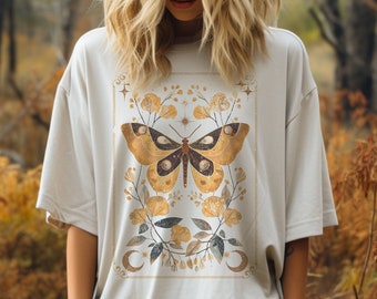May include: A white oversized t-shirt with a gold and brown butterfly graphic design. The butterfly has a celestial design with stars and moons. The shirt is worn by a person in a forest setting.