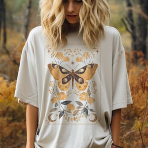 May include: A white oversized t-shirt with a gold and brown butterfly graphic design. The butterfly has a celestial design with stars and moons. The shirt is worn by a person in a forest setting.