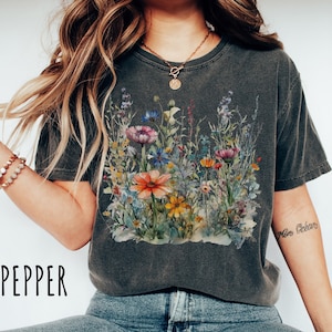 May include: A dark gray t-shirt with a watercolor floral print. The print features a variety of flowers in shades of pink, orange, yellow, and blue. The shirt has a relaxed fit and a crew neckline.