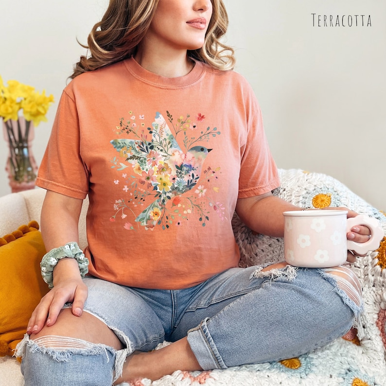 Op de afbeelding: Een terracottakleurig T-shirt met een bloemenprint met een vogel. De vogel is omringd door bloemen in tinten roze, blauw, geel en groen. Het shirt is gemaakt van een zacht, comfortabel materiaal en heeft een ontspannen pasvorm.