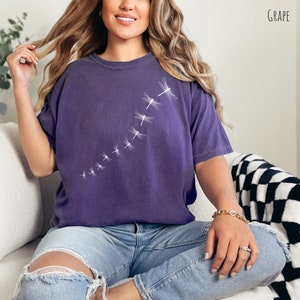 May include: A purple t-shirt with a white dragonfly design. The dragonfly design is a line of dragonflies flying in a diagonal line from the bottom left to the top right of the shirt.