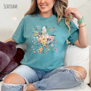 Op de afbeelding: Een turquoise t-shirt met een bloemenprint met een vogel. De vogel is omringd door roze, gele en blauwe bloemen. Het shirt wordt gedragen door iemand die op een witte bank zit.