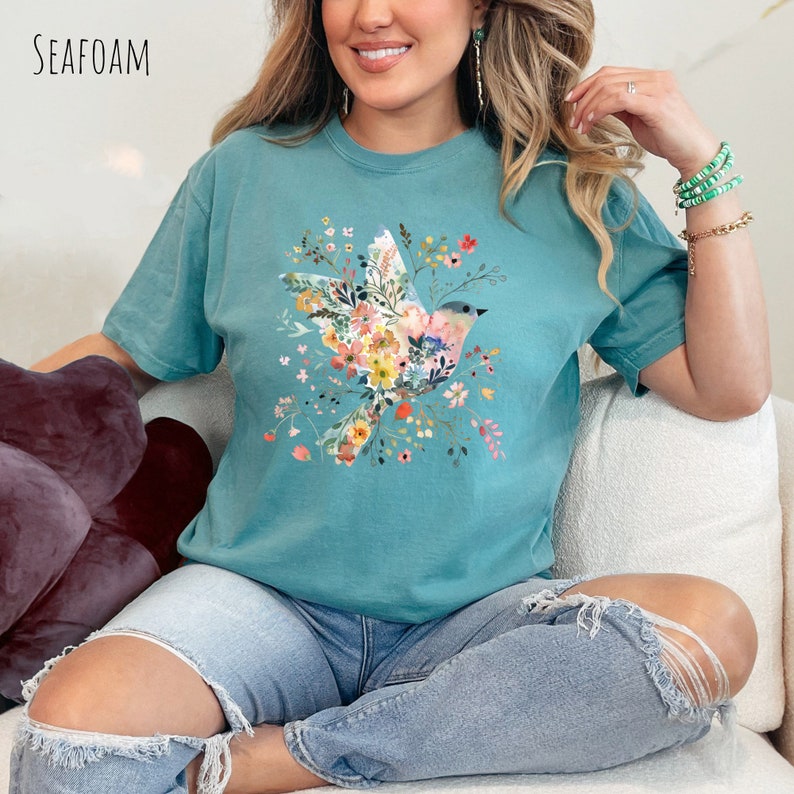 Op de afbeelding: Een turquoise t-shirt met een bloemenprint met een vogel. De vogel is omringd door roze, gele en blauwe bloemen. Het shirt wordt gedragen door iemand die op een witte bank zit.