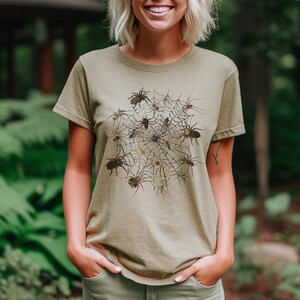 May include: A light green t-shirt featuring a graphic print of a spider's web with spiders in various poses. The spiders are brown and black and are arranged in a circle around the web.