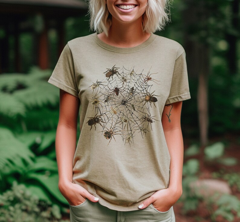 May include: A light green t-shirt featuring a graphic print of a spider's web with spiders in various poses. The spiders are brown and black and are arranged in a circle around the web.