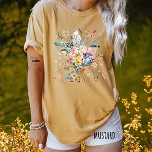 Op de afbeelding: Een mosterdgeel T-shirt met een bloemenontwerp met een vogel. De vogel is gemaakt van bloemen en heeft een blauwe kop. Het shirt heeft een ronde hals en korte mouwen.