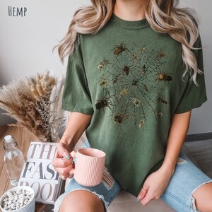 May include: A green t-shirt featuring a graphic print of a spider web with spiders. The shirt is made from hemp.