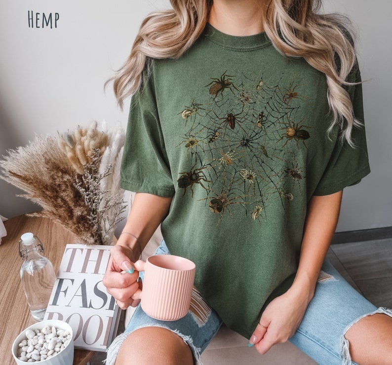 May include: A green t-shirt featuring a graphic print of a spider web with spiders. The shirt is made from hemp.