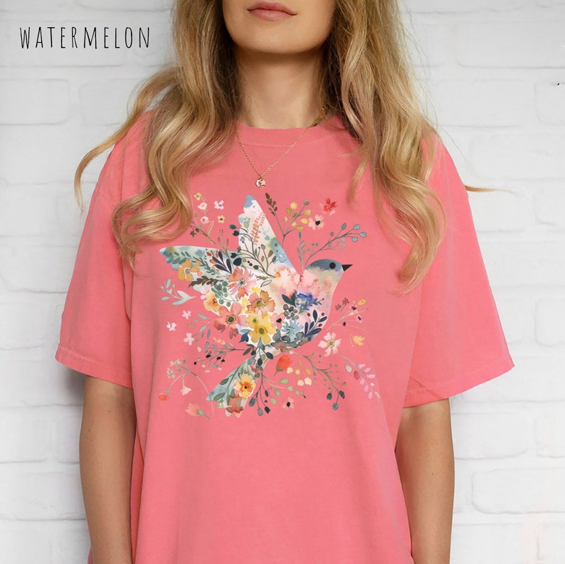 Op de afbeelding: Een roze T-shirt met een bloemenprint met een vogel. De vogel is gemaakt van bloemen en bladeren.