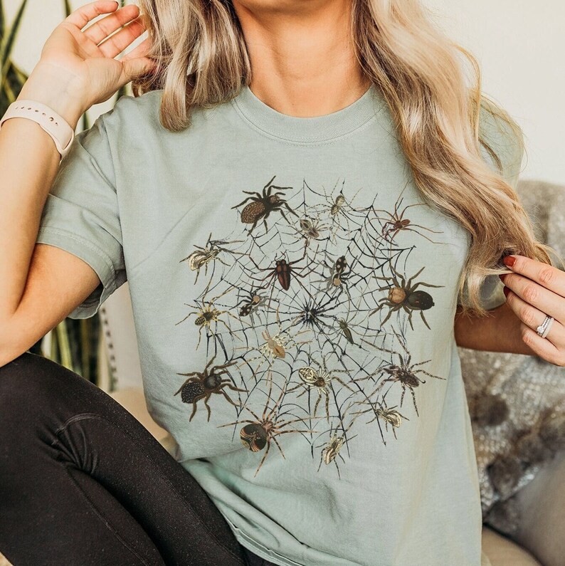 May include: A light blue t-shirt featuring a graphic print of a spider web with numerous spiders in different positions.