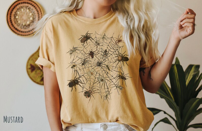 May include: A mustard yellow t-shirt featuring a graphic print of a spider web with multiple spiders of varying sizes and positions.